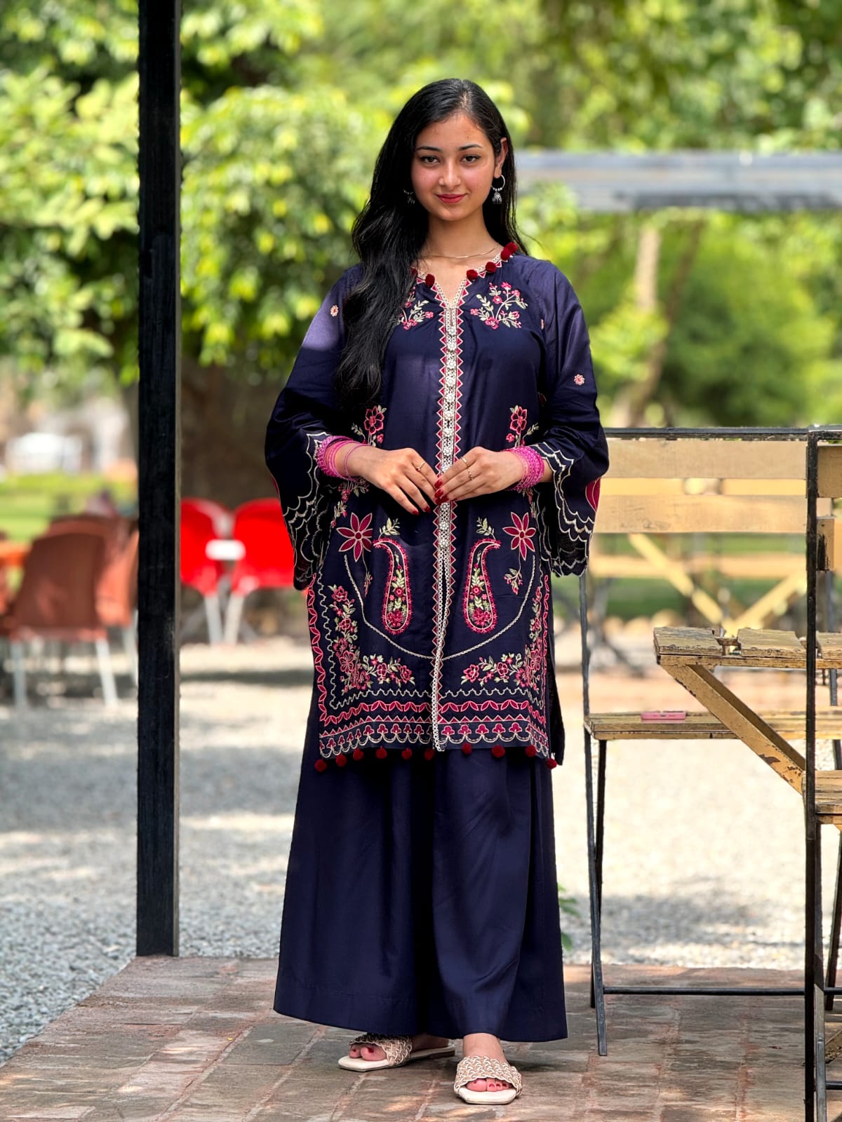 Woman wearing a dark blue embroidered traditional outfit standing outdoors with greenery in the background.